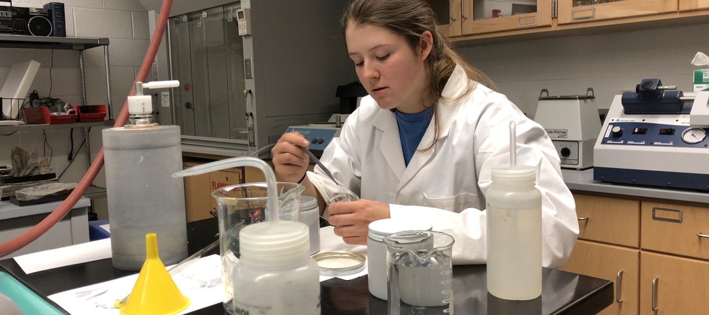 Student wearing lab coat conducts experiment in the rock preparation lab